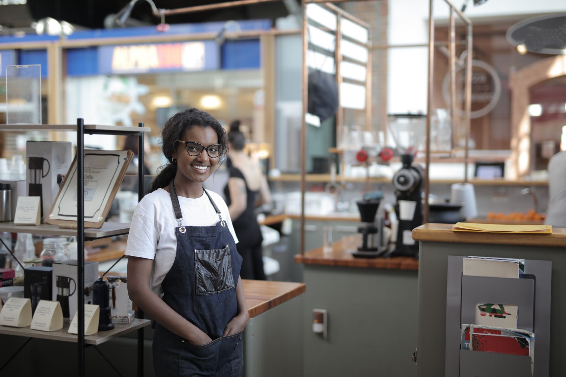 Smiling small-business owner in a denim apron standing at the counter of a coffee and homewares shop